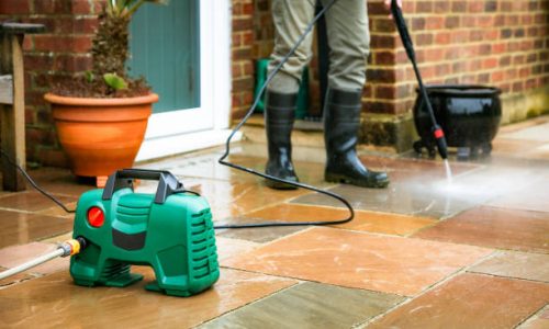 Patio Maintenance 2 A senior man in his 70s cleans his patio stone slabs using a pressure washer. The man wears a green and blue check shirt with a green gilet.